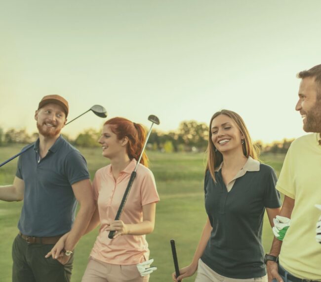 Groupe d’amis souriants marchant sur le parcours du Club de golf Val-Neigette, sacs à l’épaule.